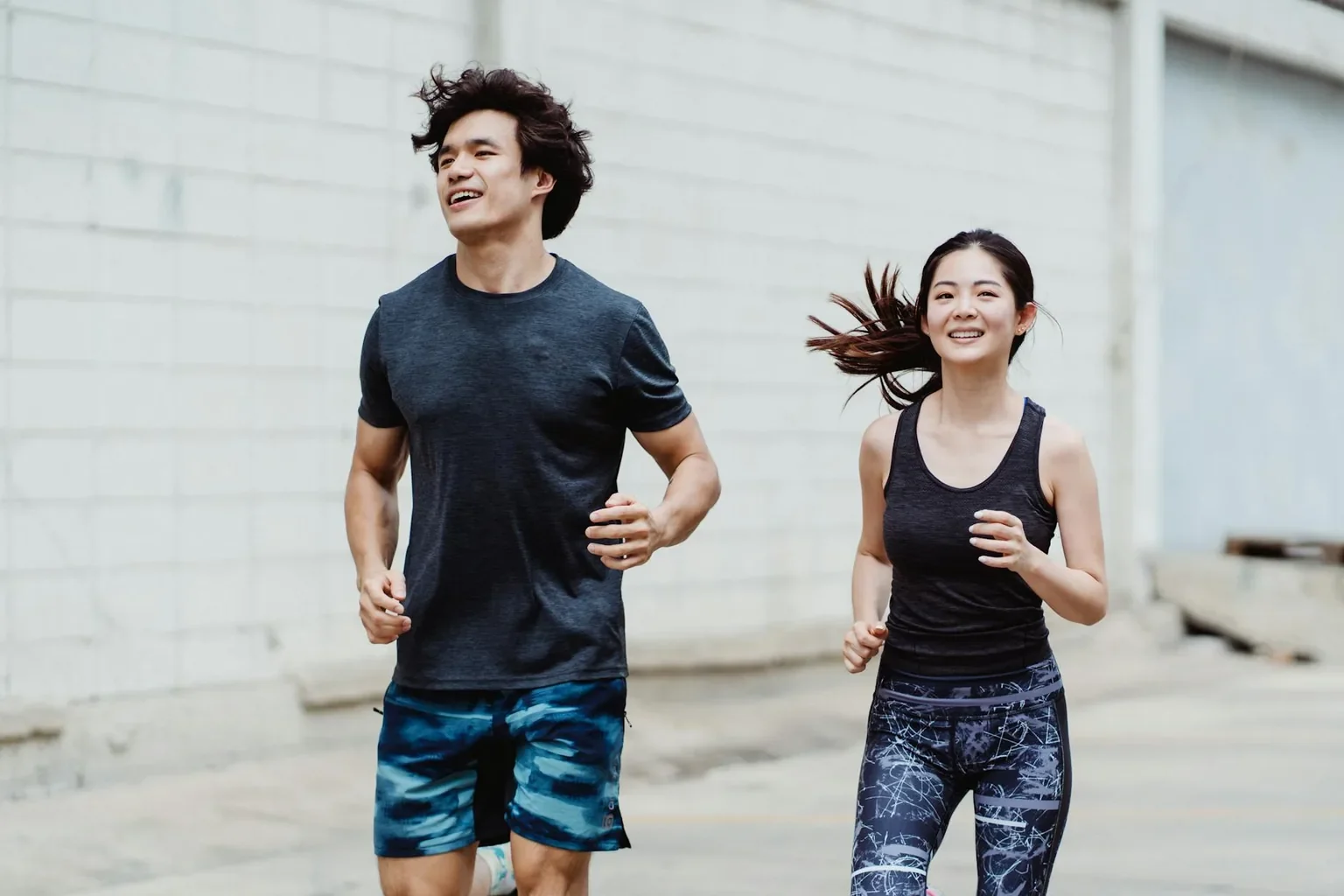 A happy couple jogging together outdoors, wearing trendy activewear and smiling energetically.