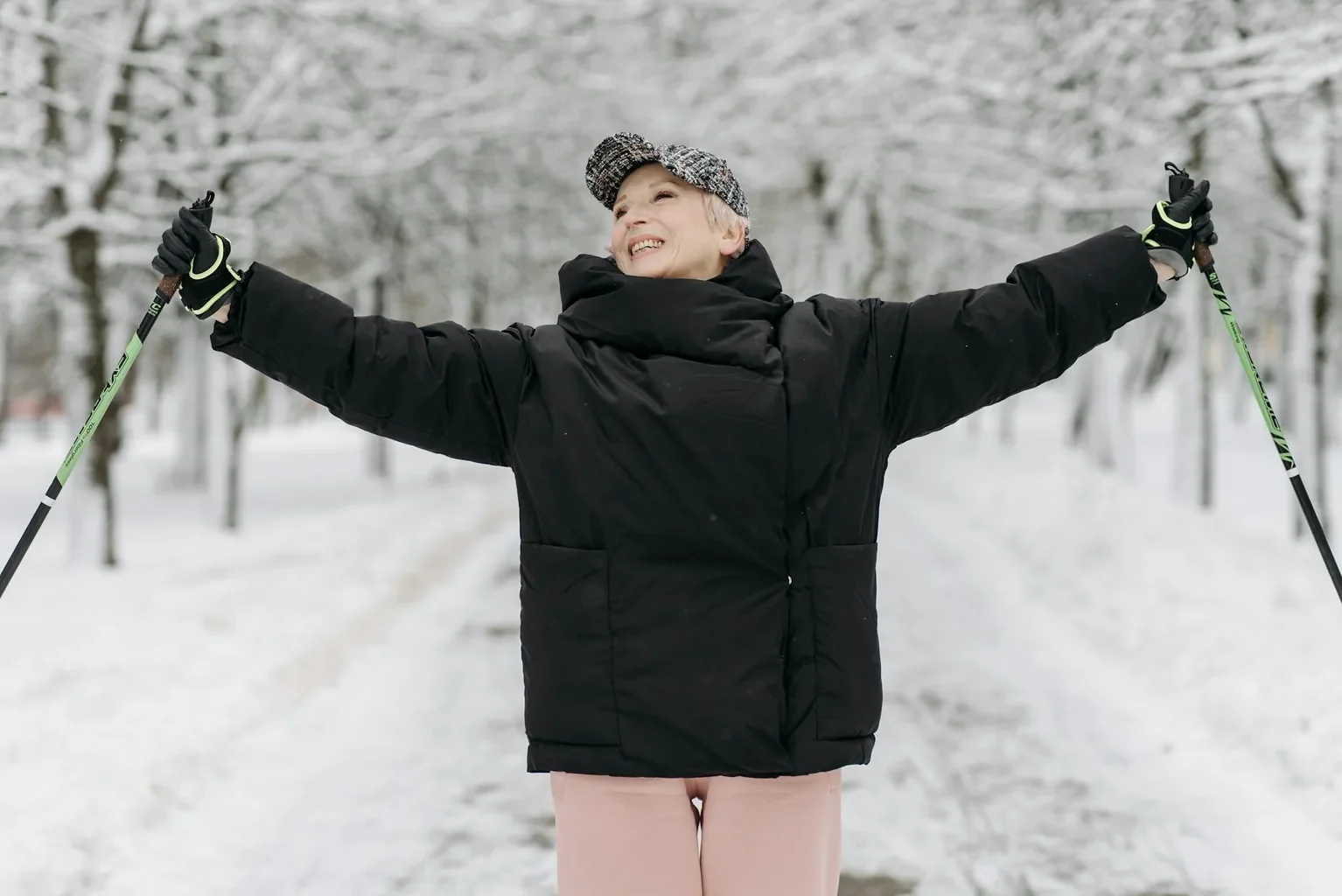 Senior woman smiling and raising arms while enjoying a winter walk with ski poles outdoors.