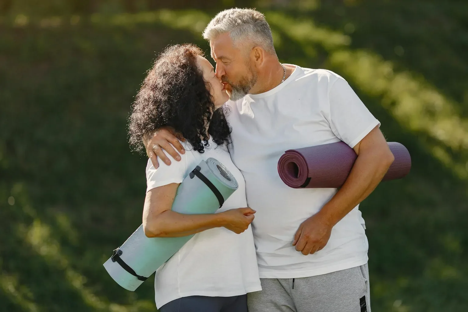 An affectionate couple embracing each other while holding yoga mats outdoors, enjoying a sunny day.