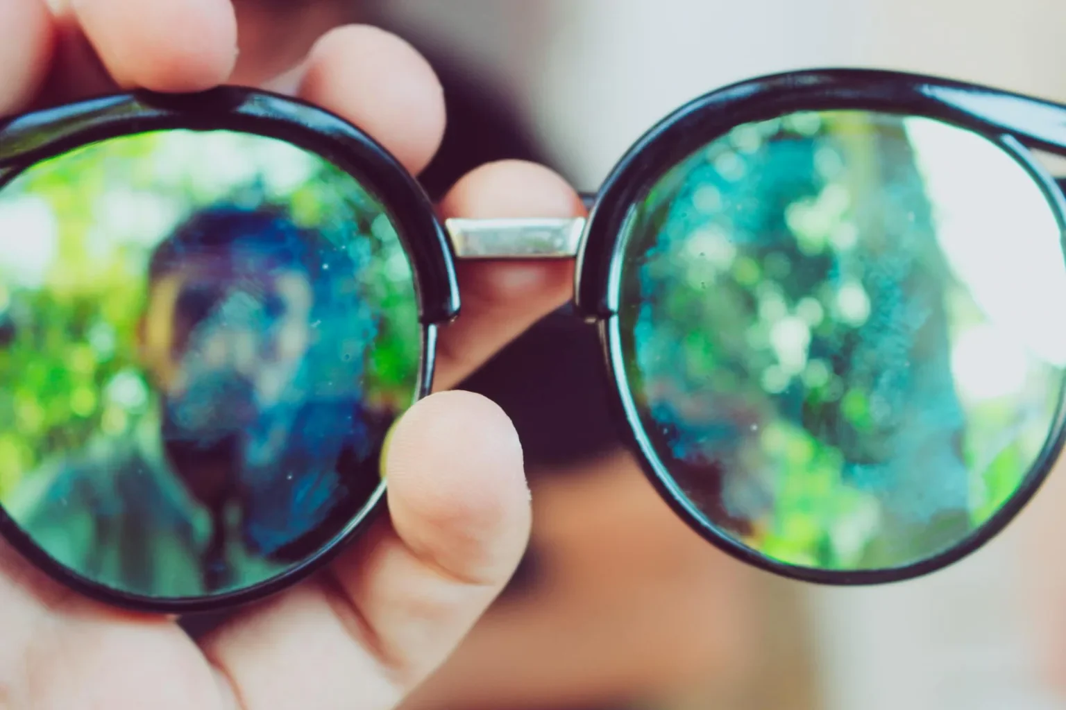 Hand holding sunglasses with nature reflection, creating an artistic blur effect.