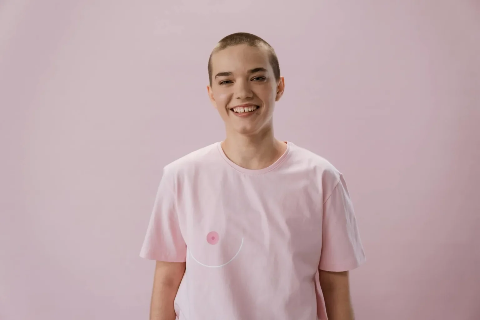 A smiling woman with a shaved head wears a pink shirt symbolizing breast cancer awareness.