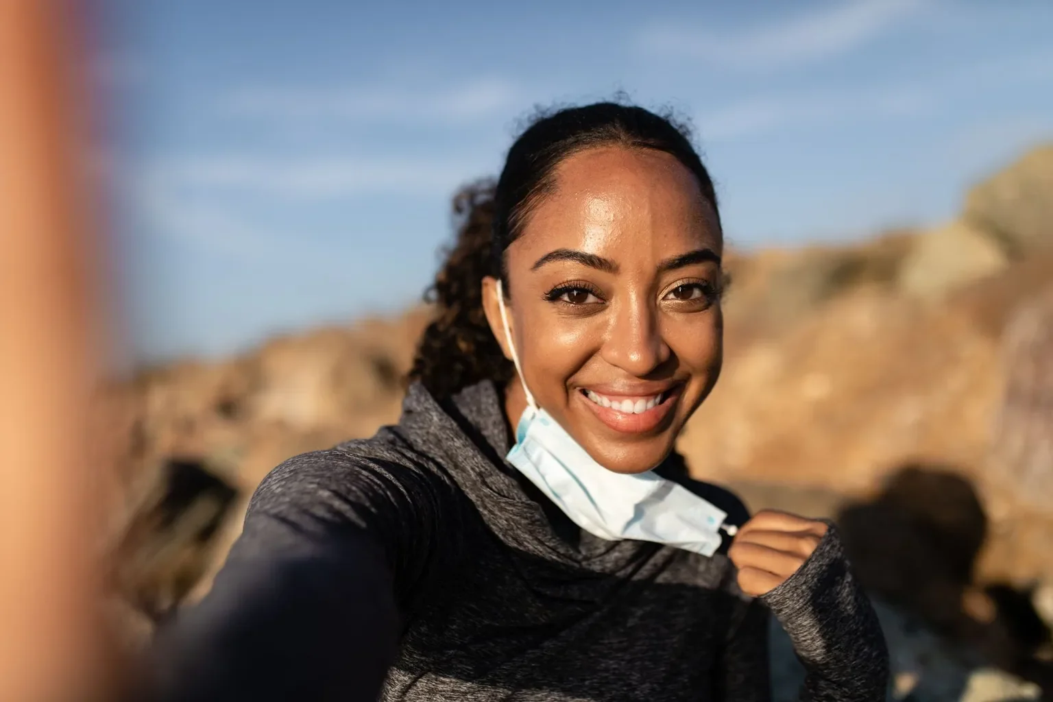 A woman smiles brightly while removing her face mask outdoors.