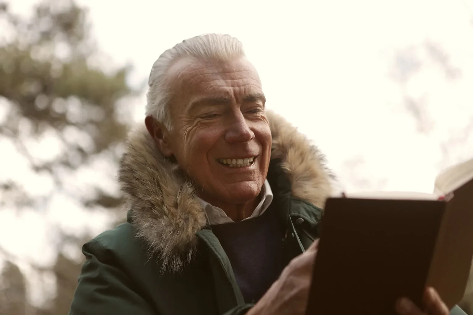 A cheerful senior man in a fur-lined coat enjoys reading a book outdoors, capturing leisure and joy.